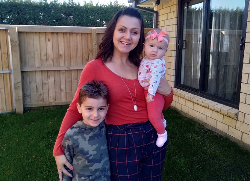 A woman with black hair, red shirt and blue trousers with red checks stands outside a house holding a young baby wearing a pink bow, pink and white patterned top and red trousers and with her arm around a boy with dark hear wearing a grey camouflage t-shirt