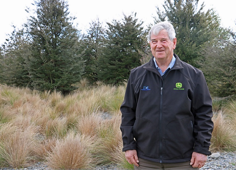 An elderly man with grey hair wearing a black windbreaker with a John Deere logo on it stands laughing in front of some small pine trees and tussock grass