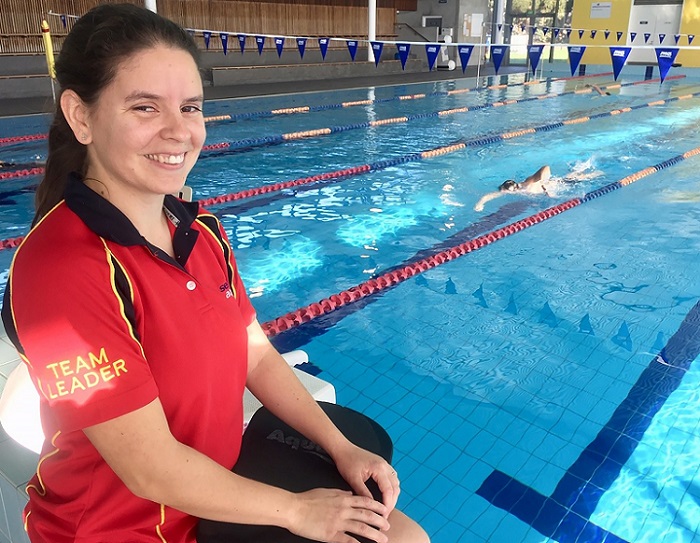 Woman with black hair wearing red and black shirt with team leader on sleeve sits on side of pool