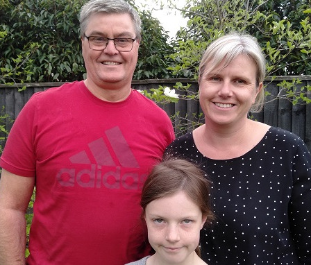 A grey haired man in glasses and a red t-shirt stands with a blonde haired woman in a white-spotted black top and a brown-haired girl 