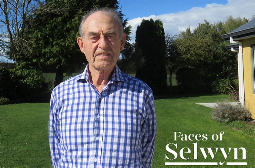 An elderly man in a blue and white checked shirt stands in a garden