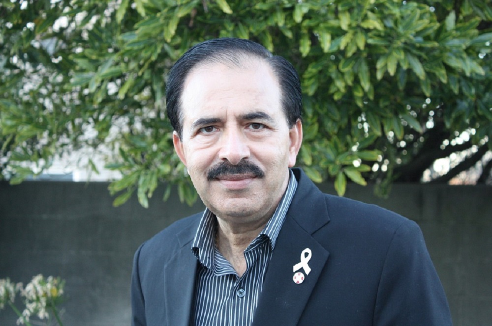 A middle-aged Middle Eastern man with receding black hair, black mustache and wearing a black suit jacket with a white ribbon lapel pin stands in front of a breeze block wall with a tree growing over the top