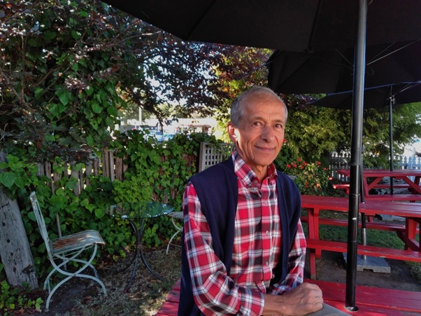 Man with receeding grey hari, red check shirt and blue gilet sits at an outdoor cafe table