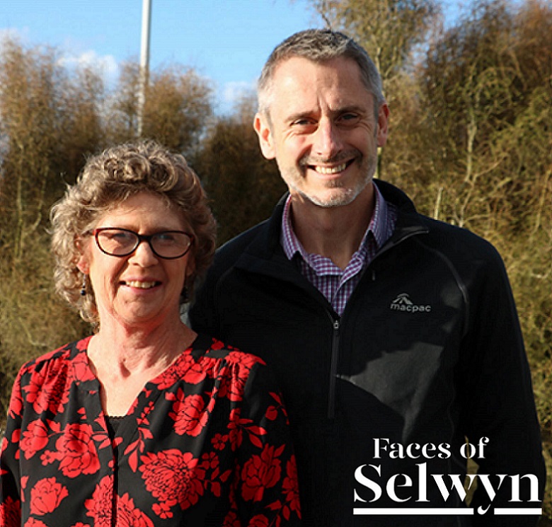 Woman with blonde curly hair and glasses in a black top with red flowers stands next to man with short grey hair and stubble wearing a blue and white checked business shirt and black mac pac zip up jacket