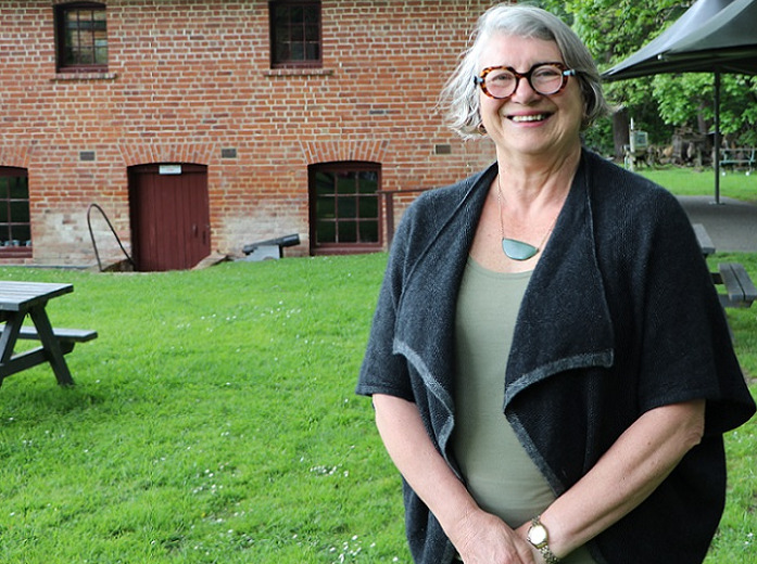 A smiling grey haired woman in round glasses, wearing a black jacket, stands on a lawn in front of a brick building