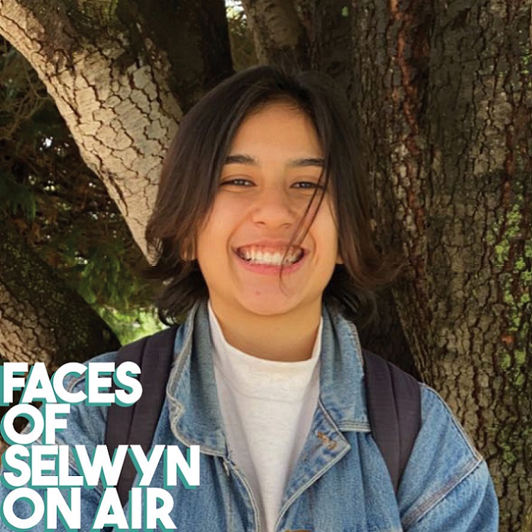A smiling person in a denim jacket and backpack standing in front of a tree