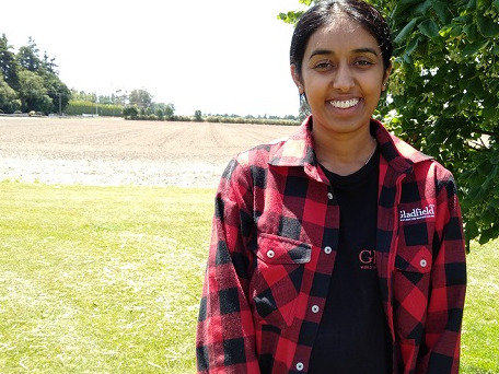 Indian woman in black and red checked shirt and black t-shirt with Gladfield Malt insignia standing on the edge of a field
