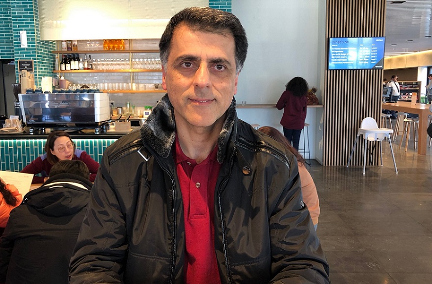 A middle eastern man in a black jacket and red shirt sits at a cafe table with the cafe counter and tables behind him