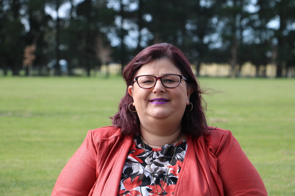A close up view of a woman with purple hair and lipstick, glasses and an orange top standing in a park