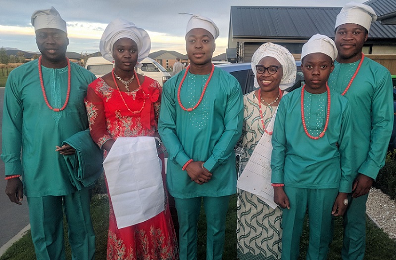 The Iposu family in traditional dress, with the four men all in green with long red necklaces and the two women in a red dress and a white and green stripped dress, both holding long white pieces of cloth and all wearing white hats. 