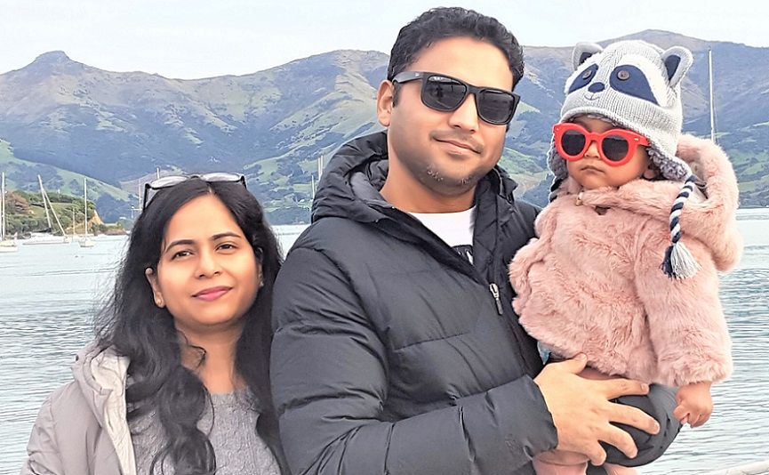 An Indian family, mum, dad and a young child in a racoon hat and sunglasses stand in front of water with a view of the port hills behind