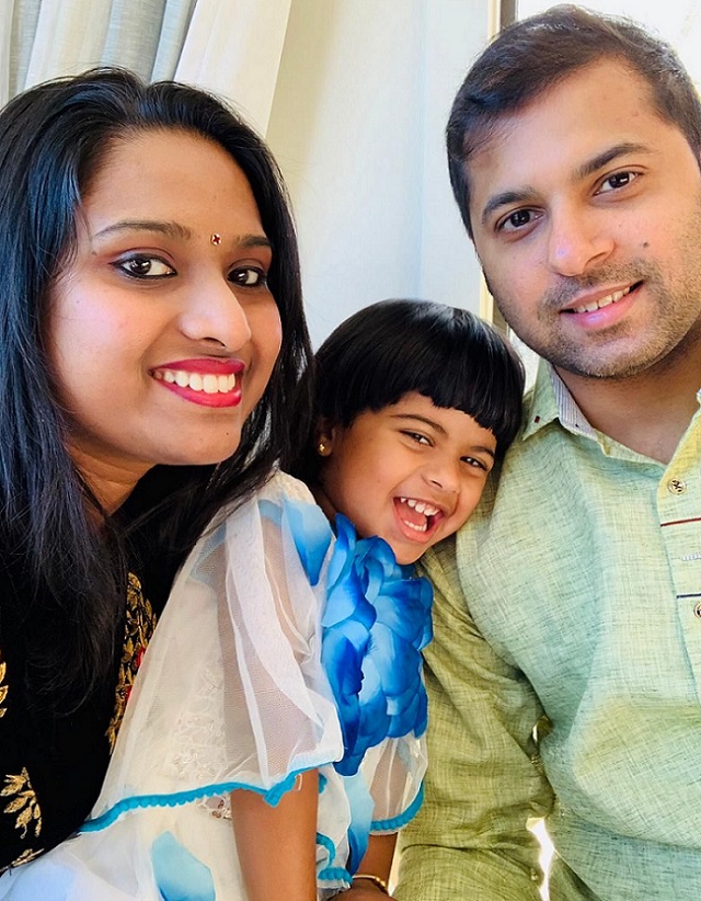 A smiling mum and dad sit leaning in close on either side of a laughing girl in a blue and white dress