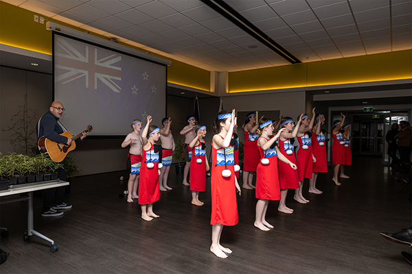 A group of primary school children perform kaphaka with guitarist and New Zealand flag in background
