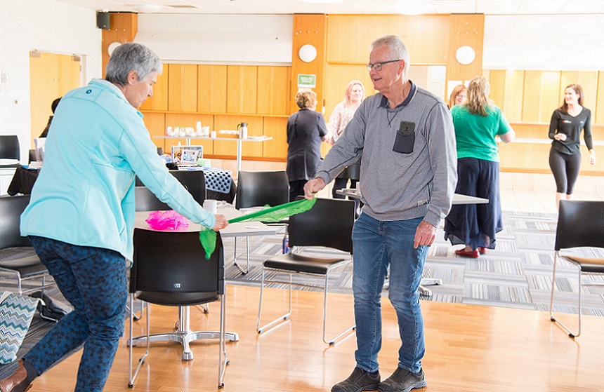 An older couple in a hall hold a green scarf taut between then while one stands on one foot