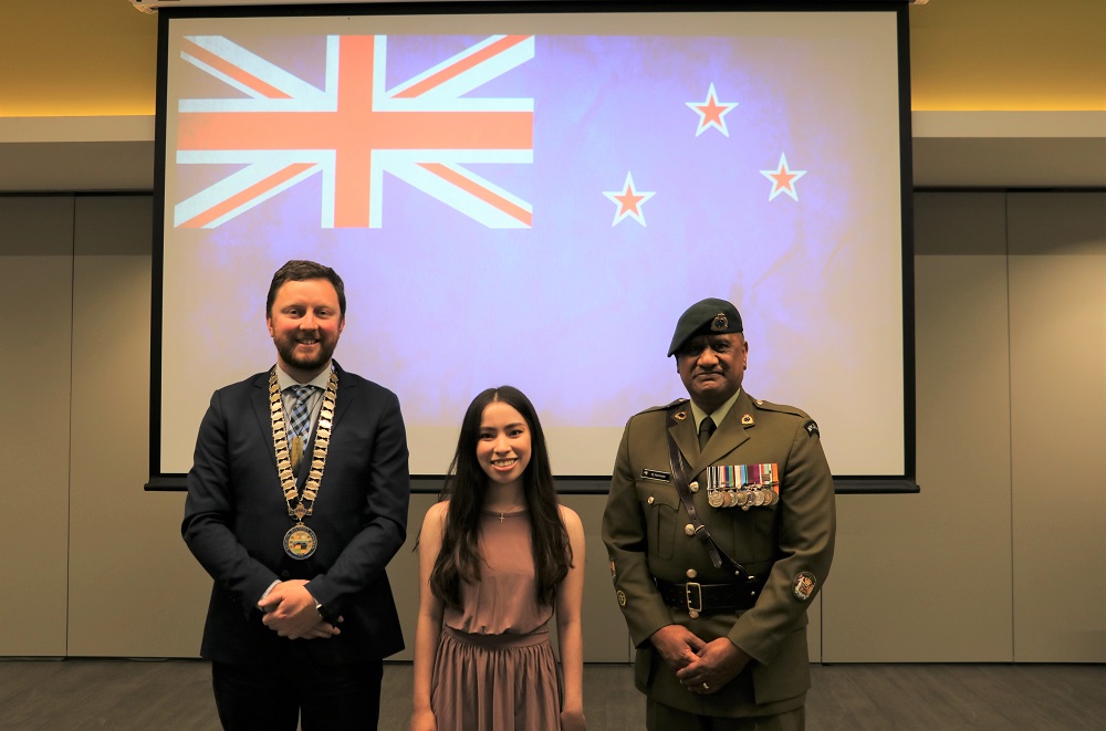 Mayor Sam Brougton in a blue suit and wearing his mayoral chains stands with a shorter woman in a grey dress and a Maori man in NZ Army dress uniform with a New Zealand flag on a scree behind them