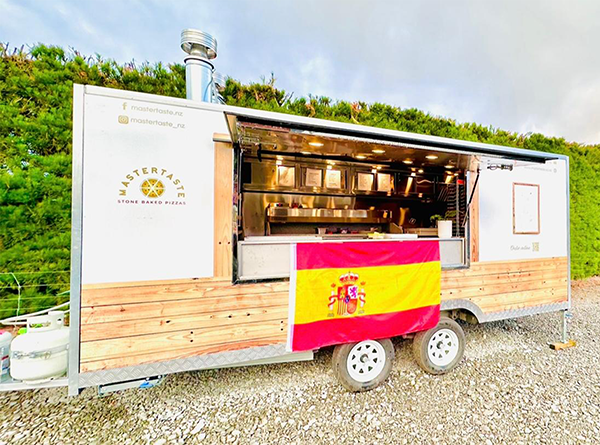 Food truck with Spanish flag in front of green hedge on gravel