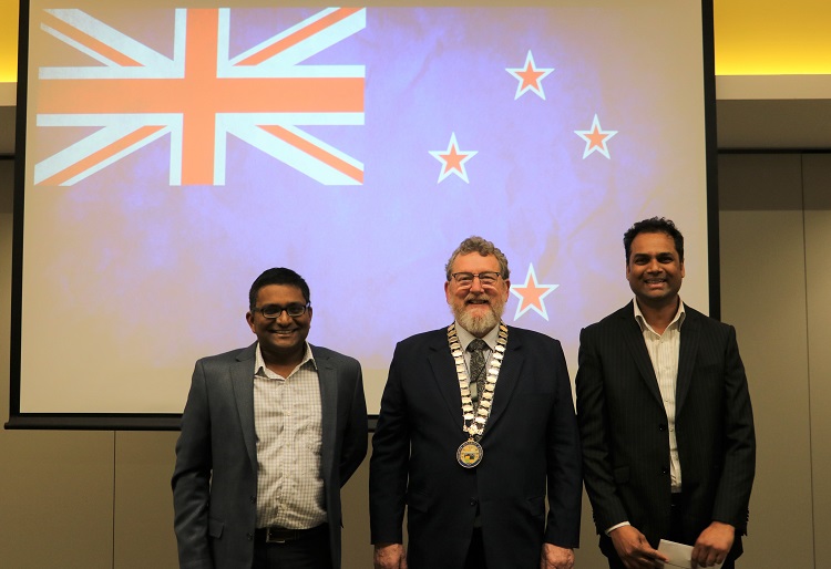 Two Indian men in suits stand either side of Deputy Mayor Malcolm Lyall with a New Zealand flag projected onto a screen behind
