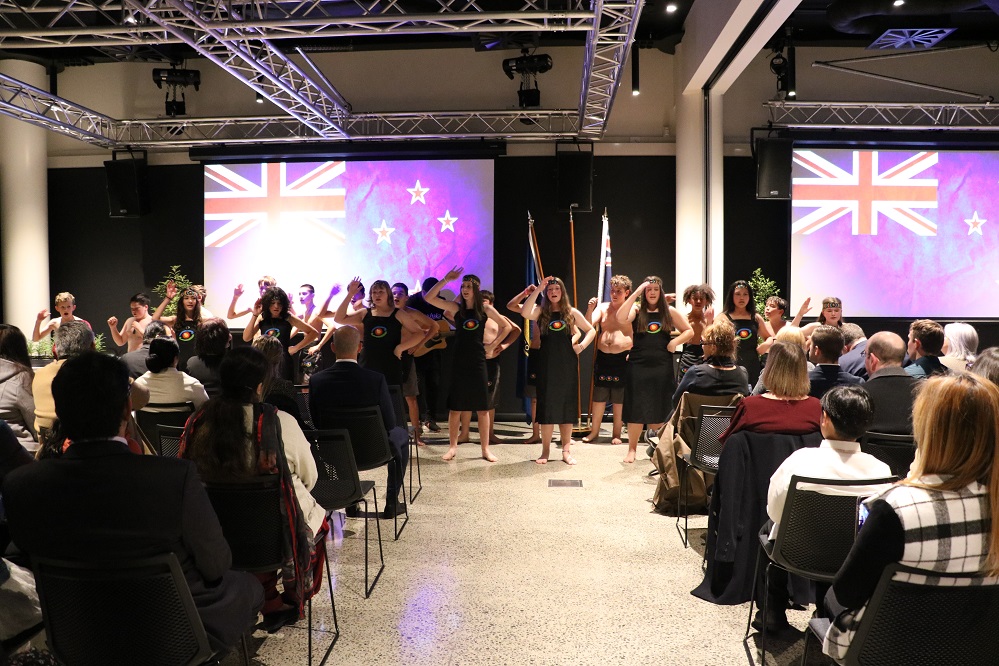 A seated crowd watch a school kapa haka group performing in front of screens with the New Zealand flag