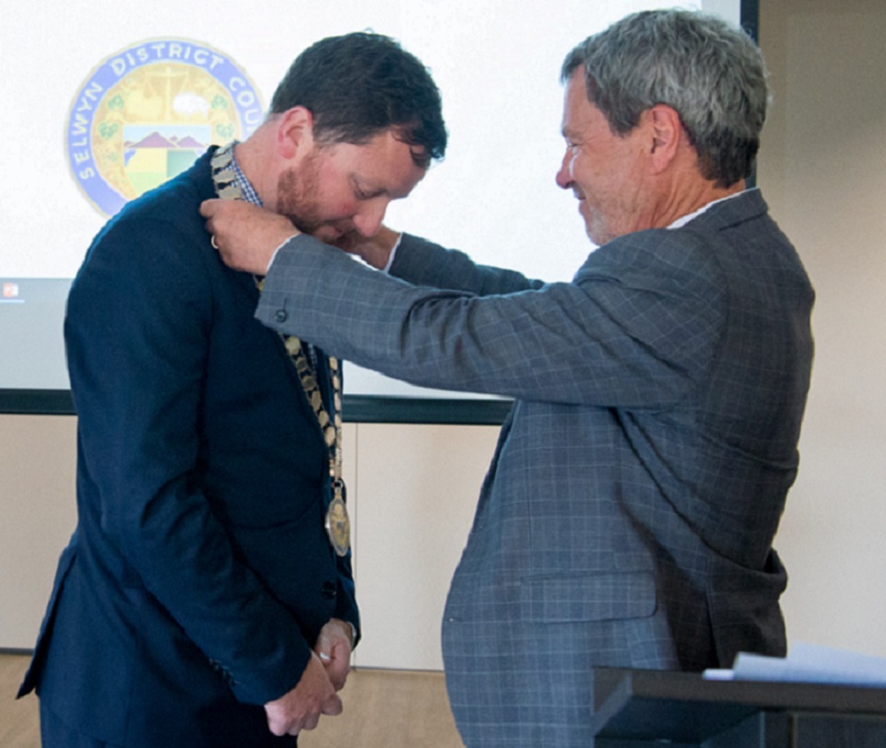 Mayor Sam Broughton, in a blue suit, with brown hair and beard, has his neck bowed as Council Chief Executive David Ward, in a grey suit, with grey hair, puts the Mayoral chains on him