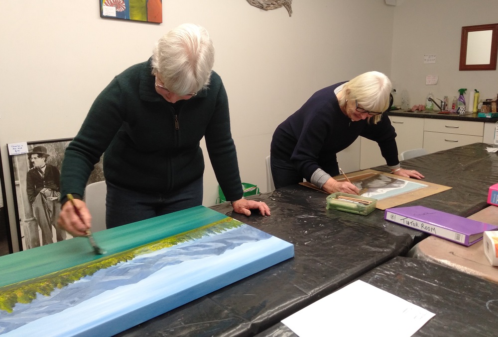 Two women paint landscape paintings on a long bench