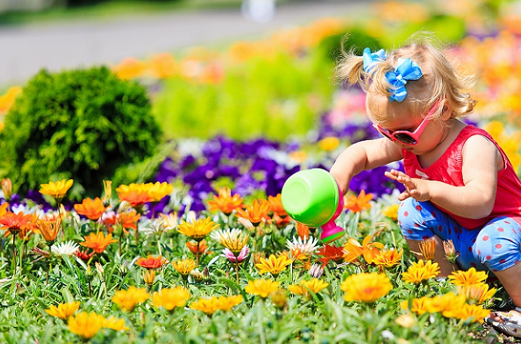 A toddler in sunglasses watering a brightly coloured flower bed