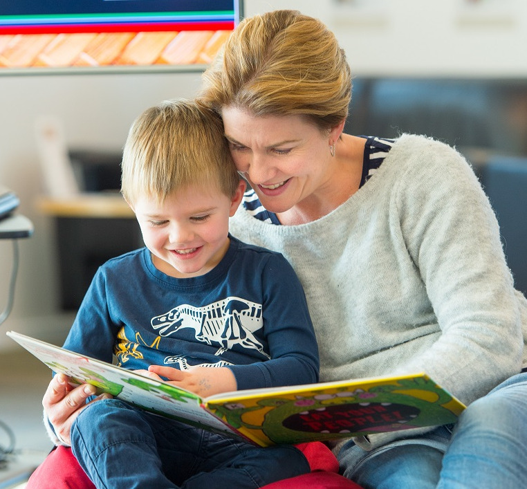 A mother and a young boy in a dinosaur t-shirt sit reading a book together on a red bean bag