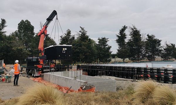 A crane lowers a large black container like box into a concrete housing watched by a man in hard hat and high vis jacket