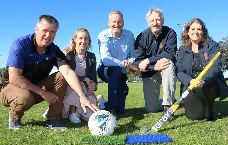 Five people, one holding a football, one holding a hockey club, crouch on a grass field behind two pieces of artificial turf