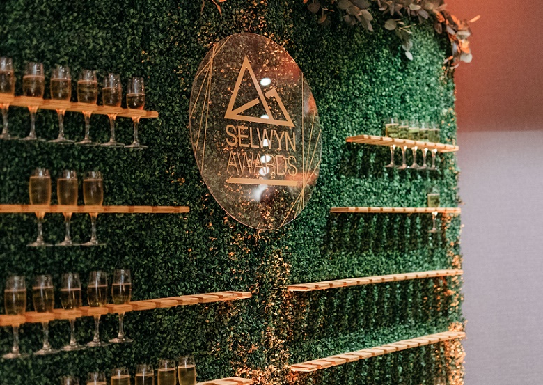 A green textured wall, with shelves on it holding glasses and a circular decorated bronze coloured sign in the middle saying Selwyn Awards