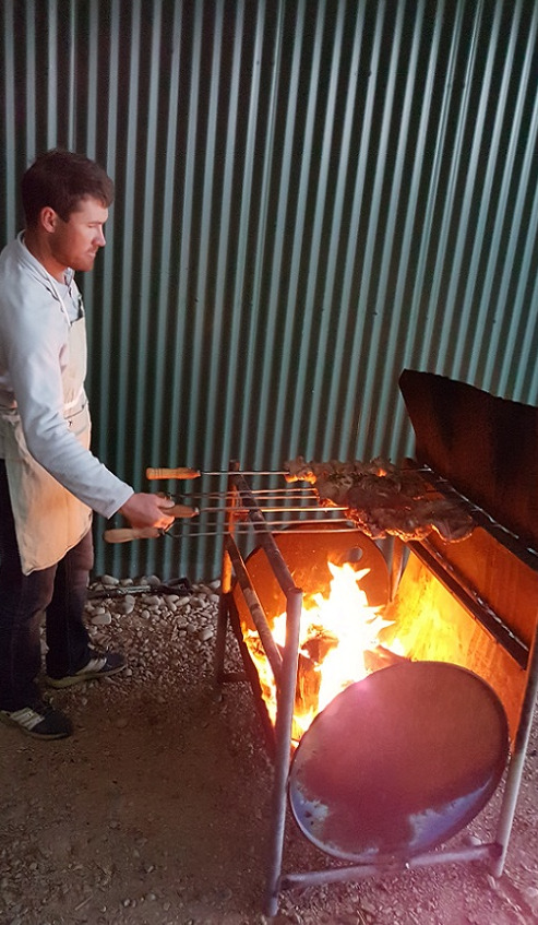 A man in white shirt and apron barbecuing meat on long skewers over a half drum barbecue