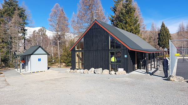 Black wooden community hall with rocks outside people standing around with trees and snow on mountains in background
