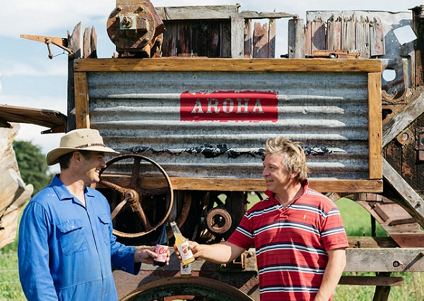 two men chink bottles in front of a corrugated iron sign with the word Aroha stenciled on it 