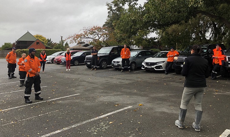 A group of men and women in orange jackets spaced through a car park some standing next to cars, listening to a woman with back to camera giving instructions