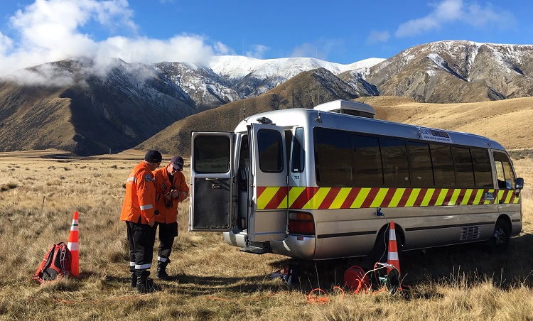 Two people in orange high visibility jackets stand behind a silver van with a red a yellow band along the side under the window line. The van is parked in a field of brown tussock at the foot of some mountains with a light dusting of snow on them