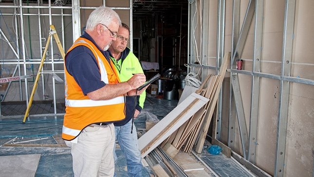 Two men in high visibility jackets stand in a half built room looking at a tablet computer which one man is writing on