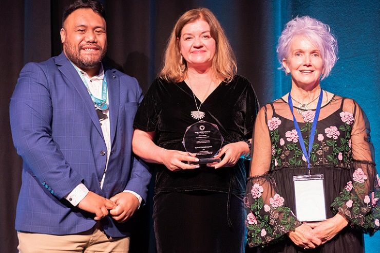 A Pasifika man and two pakeha women standing on an awards stage with the woman in the middle holding a trophy