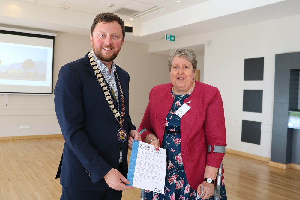 Mayor Sam Broughton wearing a blue suit and his mayoral chains and an older lady with grey hair, wearing a pink jacket, a blue dress with pink flower drawings on it and resting on crutches both hold up the accessibility charter