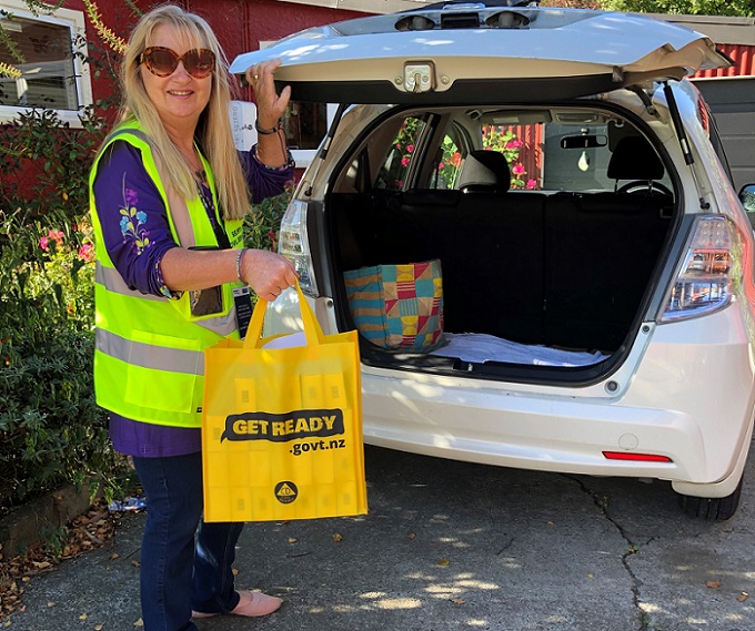 A woman in a high viz yellow jacket standing next to the open boot of a car, holding a yellow bag contained RAT kits