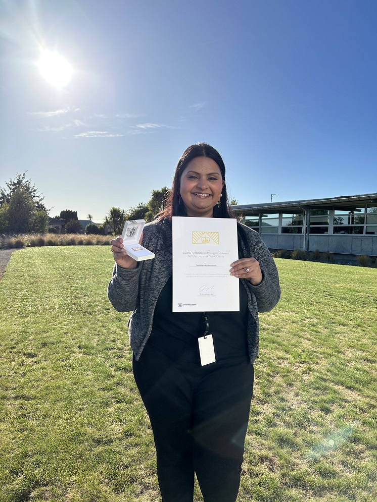 image of smiling woman outside in the sunshine holding up a certificate and box with a badge pin in it