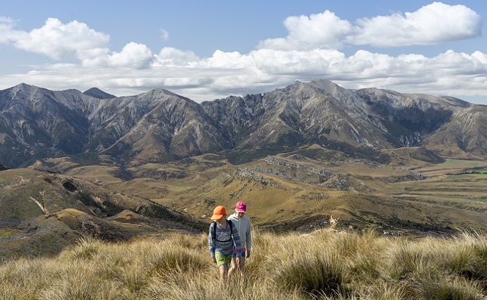 Two children, in brightly coloured caps climb a hillside with a view of valley and mountains behind
