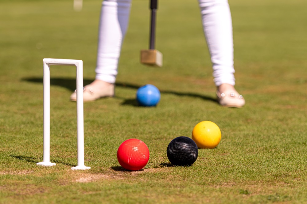 image showing a croquet lawn with three coloured ball red, black and yellow in the foreground and the feet and legs of a person getting ready to hit a blue ball with their mallet blurry in the background