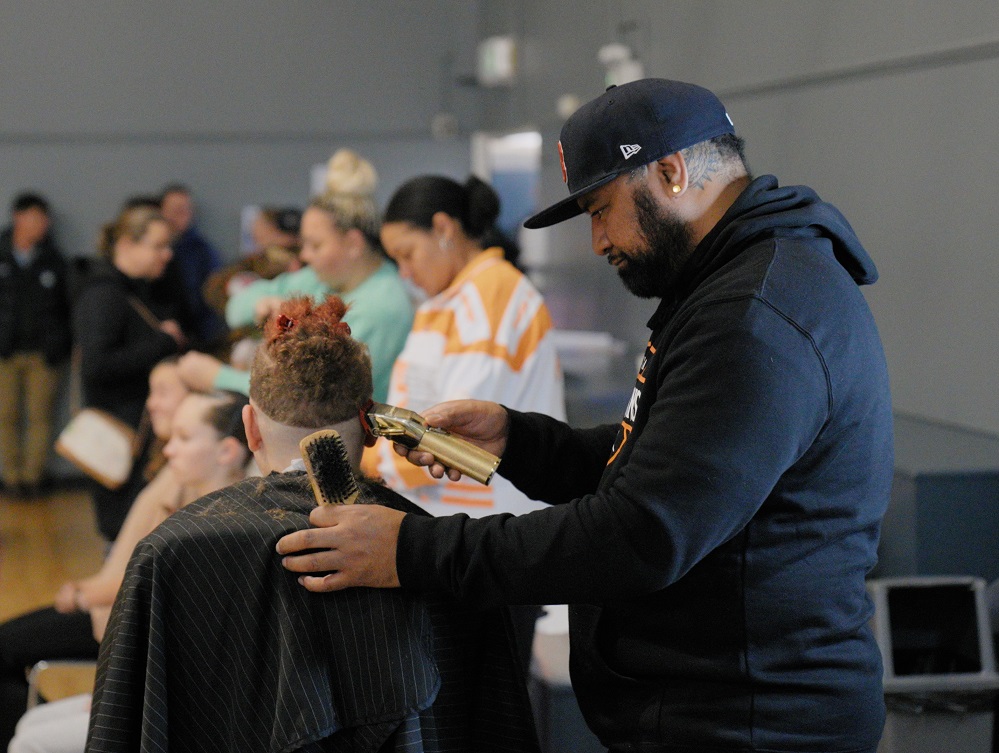 A barber in a baseball cap cuts the hair of a person facing away from the camera