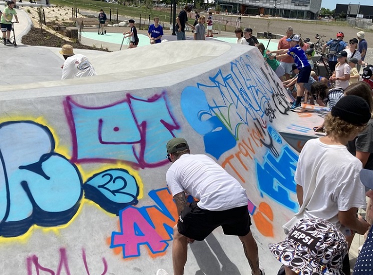 A man bent over spray painting a concrete skate ramp with street art while a crowd watches