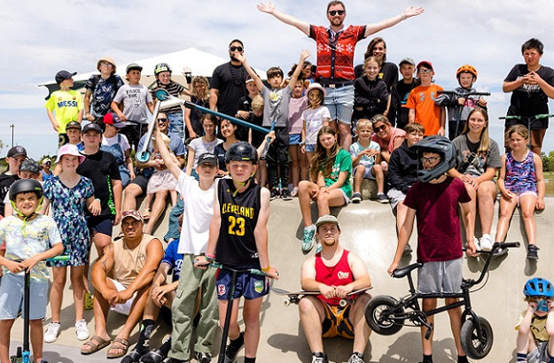 A crowd of children with scooters, skateboards and bikes together with Mayor Sam in a Christmas jumper all with their arms in the air celebrating