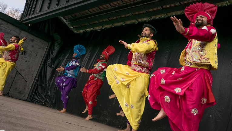 Five men in brightly coloured traditional Indian outfits in red, yellow, pink, purple and blue dance on a stage