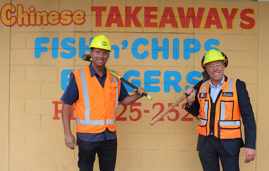 Council Grant Millar and Group Manager Property Douglas Marshall stand in front of the Chinese takeaway building in hard hats, hi-vis vests and goggles, holding sledgehammers