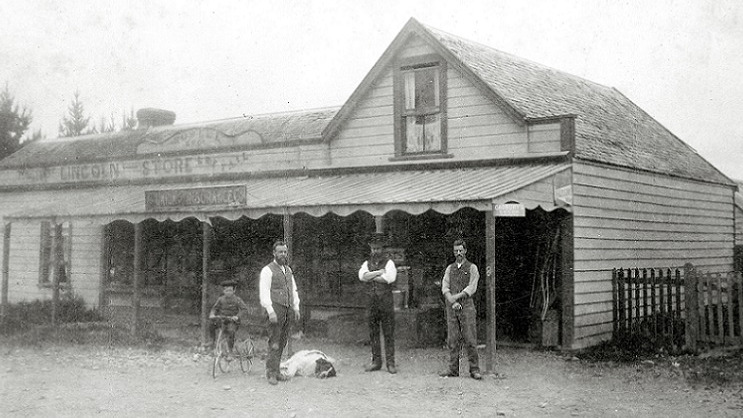 A faded black and white photograph of a child on a bicycle and three men in suits standing in front of a wooden building labeled Lincoln Store. A large white dog with a brown haired head lies on the ground at one of the men's feet