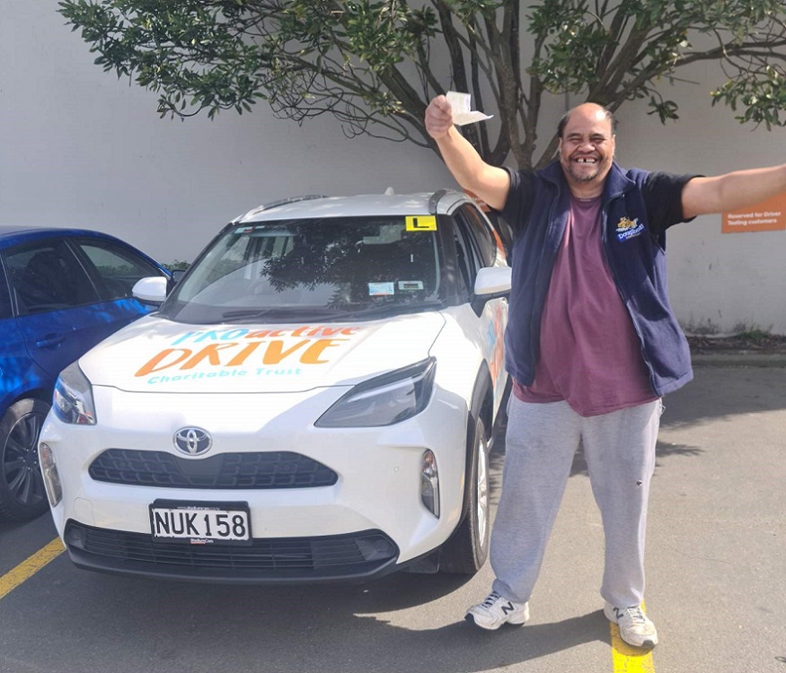 A man in track pants, a purple shirt and blue jersey smiles holding arms up in triumph in front of a Proactive Drive Charitable Trust car