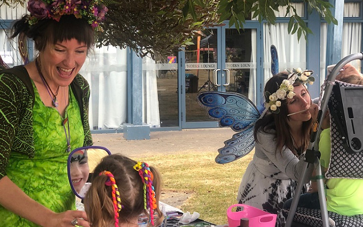 A woman dressed as a fairy in bright green smiles as she holds up a mirror to a young girl getting face painted while a woman dressed as a blue fairy paints another child's face