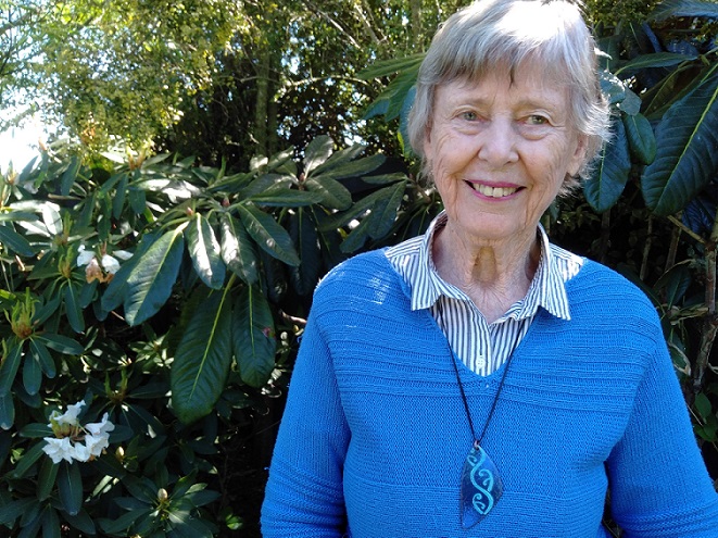 Older woman with grey hair, a blue jersey and blue necklace with traditional Maori patterning stands in front of some trees